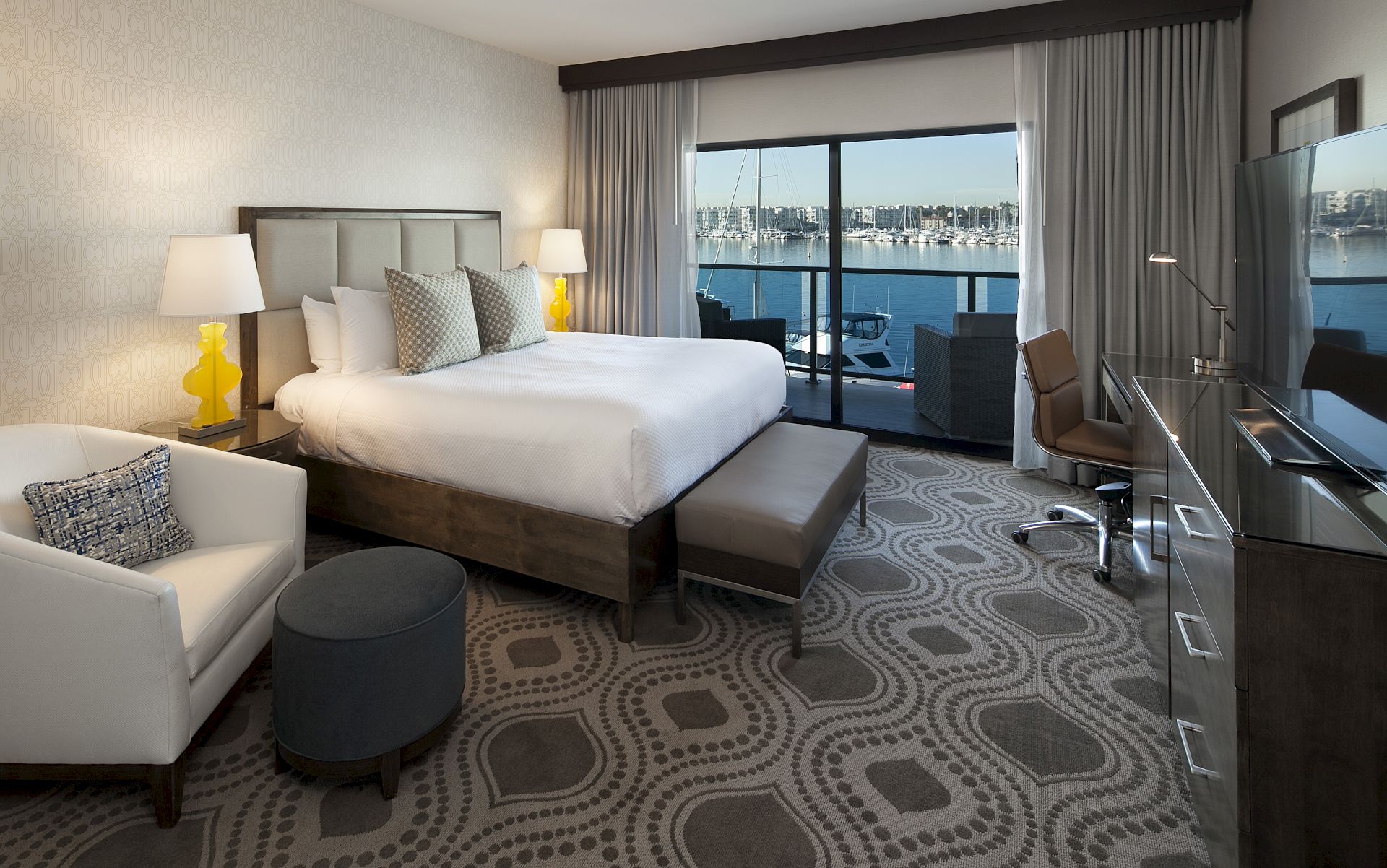 A modern hotel bedroom with a king bed, lamps, patterned carpet, and large window overlooking a marina and boats.