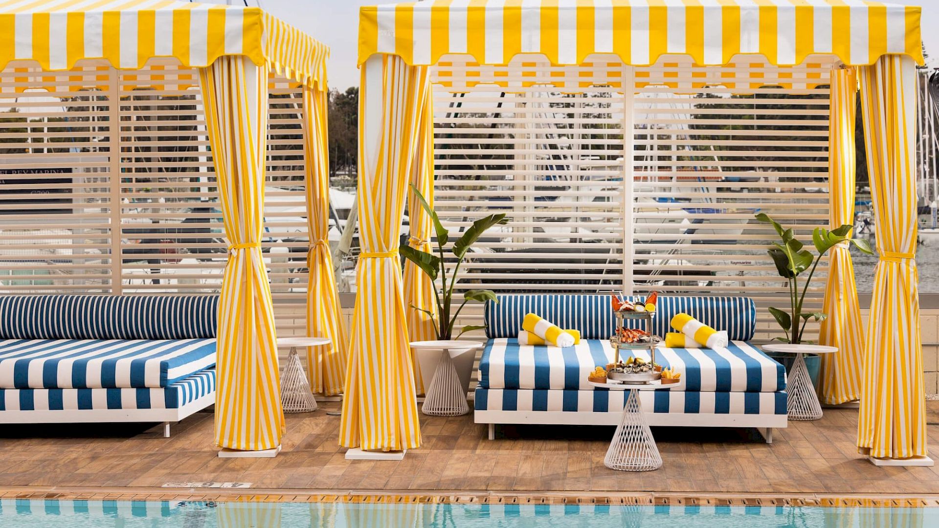 Two striped cabanas with yellow-and-white curtains by a pool, blue-and-white cushions, potted plants, and lounge chairs.
