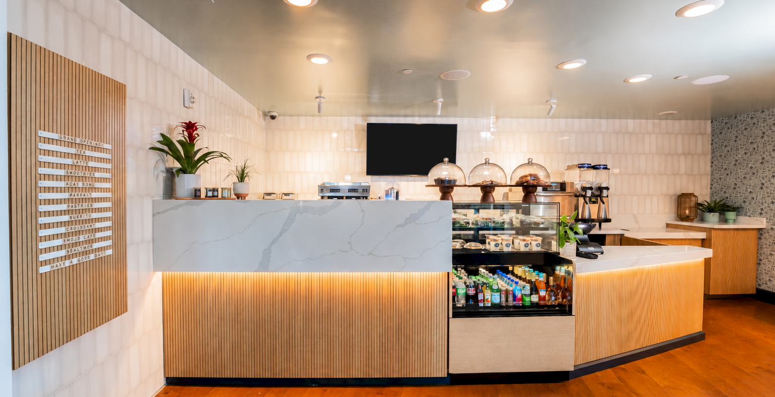A bright caf&eacute; counter with barista machines, drink dispensers, plants, and a menu board on the left.