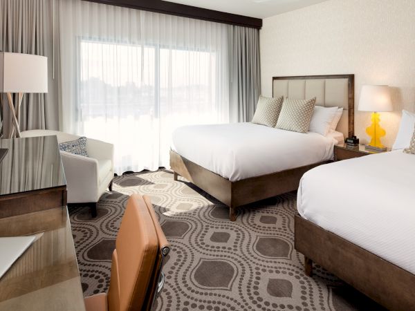 A modern hotel room with two queen beds, a desk, a chair, a dresser, a lamp, and a large window with sheer curtains, all in neutral tones.