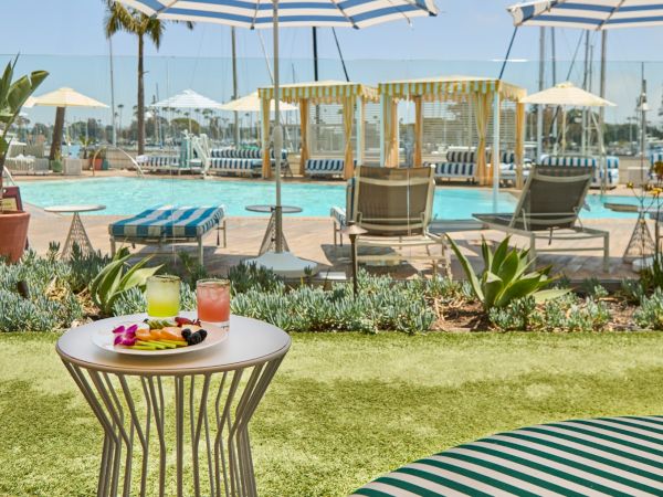 A poolside scene with lounge chairs, umbrellas, and a small table set with drinks and snacks in a sunny resort garden.