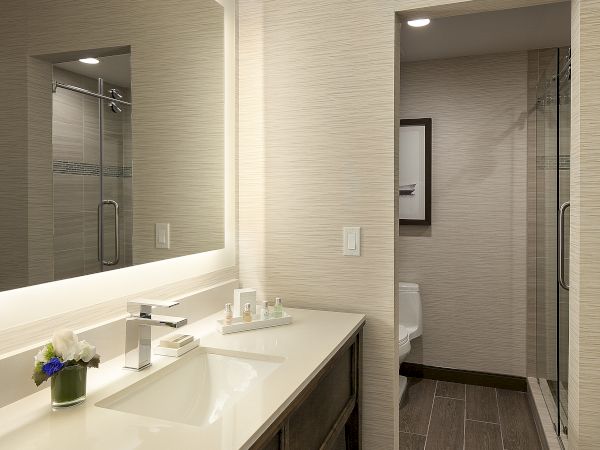 A modern bathroom with a long double sink, large mirror, subtle beige tiles, and a separate shower area visible through the doorway.