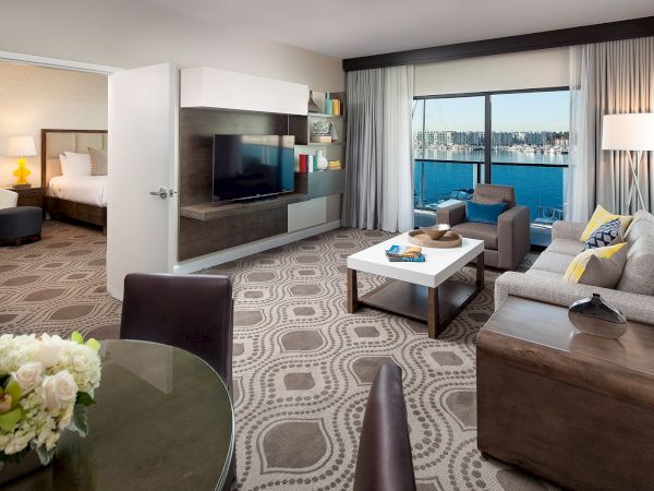 A modern hotel suite with a living area, adjoining bedroom, and a balcony view; stylish sofas, round coffee table, TV wall, and patterned carpet.