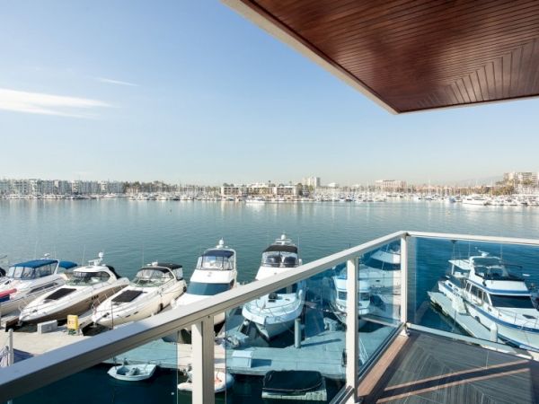 A marina view from a balcony: calm water, several boats docked, clear sky, distant buildings along the shoreline, wooden balcony railing in foreground.