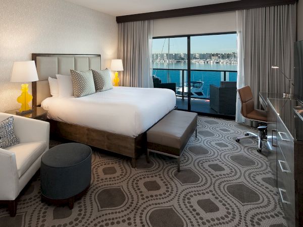 A modern hotel room with a king bed, seating area, ottoman, desk, and a balcony with a city/port view.