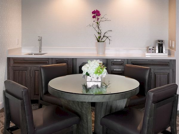 A modern kitchenette with a round glass table, six dark chairs, a small flower centerpiece, and a minimalist white-tiled backsplash with subtle lighting.