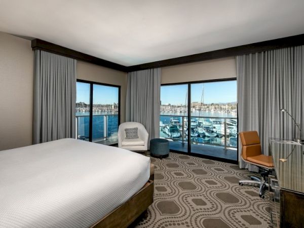 A modern hotel room with a large window view of a marina, a king bed, seating area, desk, and neutral decor, overlooking blue water.
