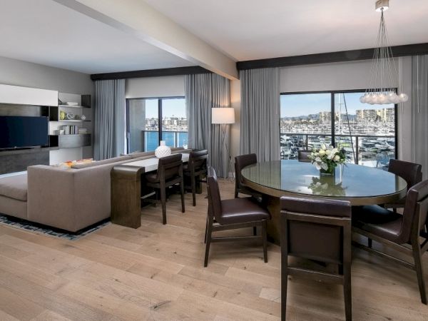 A modern, airy living-dining area with a round dining table, dark chairs, a beige sofa, a mounted TV, and floor-to-ceiling windows offering city views.