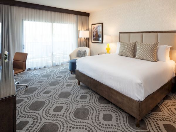 A modern hotel room with a large bed, nightstands, lamps, seating area, and a patterned carpet, plus a balcony view. Top it at 140 characters, always ending the sentence.