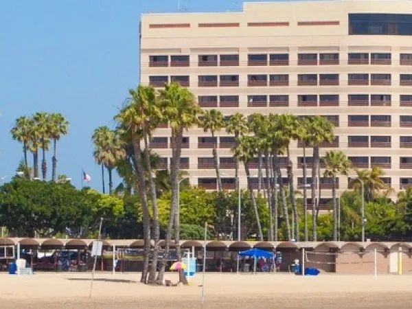 A beach with sandy shore, palm trees, a few people, and a multi-story beige building in the background, under a clear blue sky.