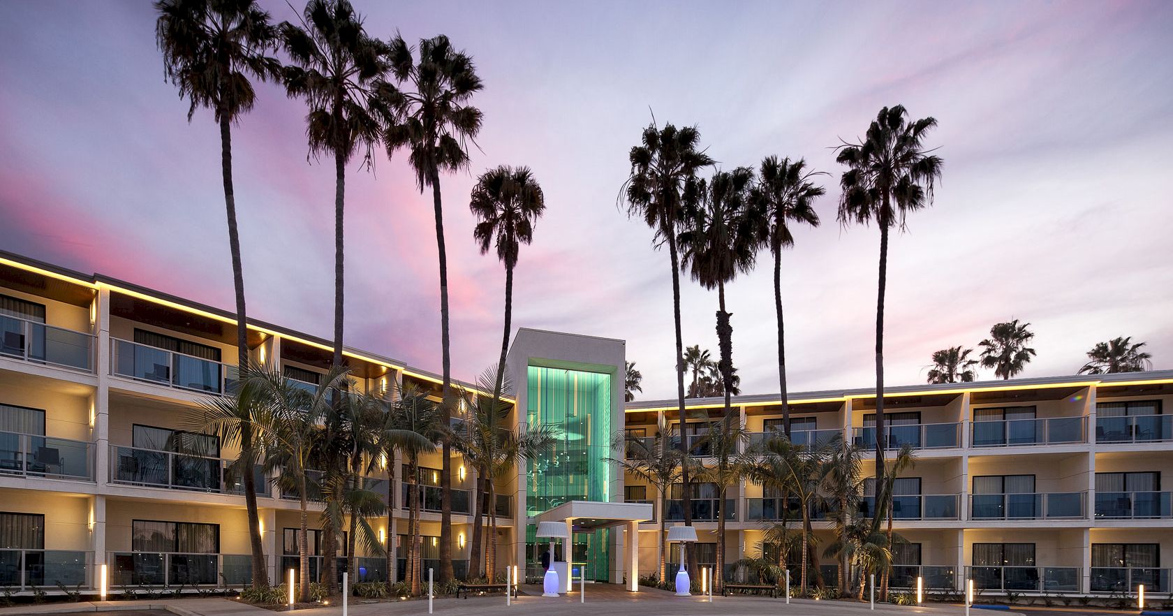 A mid-century motel-style building with multiple floors, tall palm trees, and a glowing green glass entrance at dusk, under a pink-purple sky.
