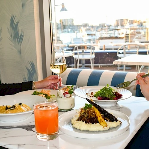 Two people dine at a bright caf&eacute; by the window, sharing drinks and plates of pasta and salad on a white-table setting.