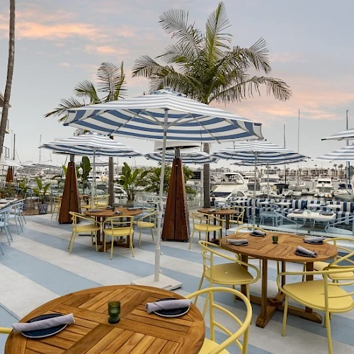 A sunny beachside cafe with wooden tables, yellow chairs, blue-striped umbrellas, and palm trees, ready for diners at sunset.