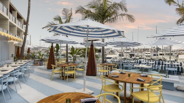 A sunny beachside cafe with wooden tables, yellow chairs, blue-striped umbrellas, and palm trees, ready for diners at sunset.