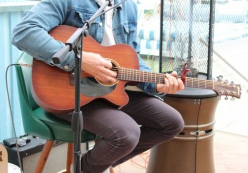 A person plays an acoustic guitar while sitting on a green chair, with a drum beside them and a mic stand nearby.