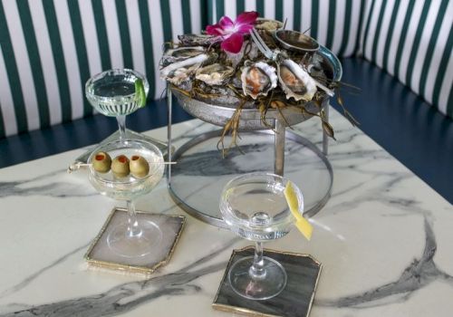 A chic coastal setup with a tiered shell centerpiece, oysters on ice, a champagne coupe with a lemon twist, and mini olives on a skewered tray, all atop marble surfaces.