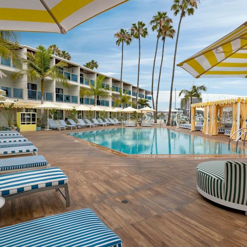 A fancy poolside resort with blue-striped loungers, palm trees, cabanas, and a bright yellow/white cabana area by the sea, sunny and relaxing.