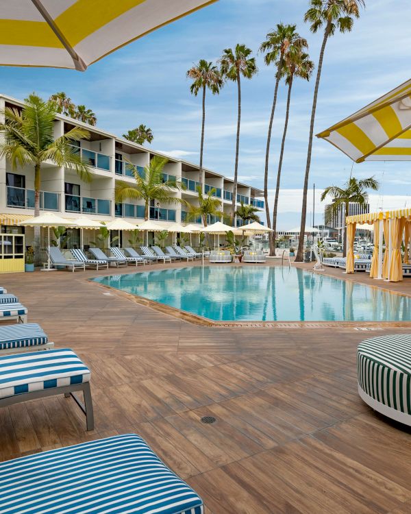 A fancy poolside resort with blue-striped loungers, palm trees, cabanas, and a bright yellow/white cabana area by the sea, sunny and relaxing.