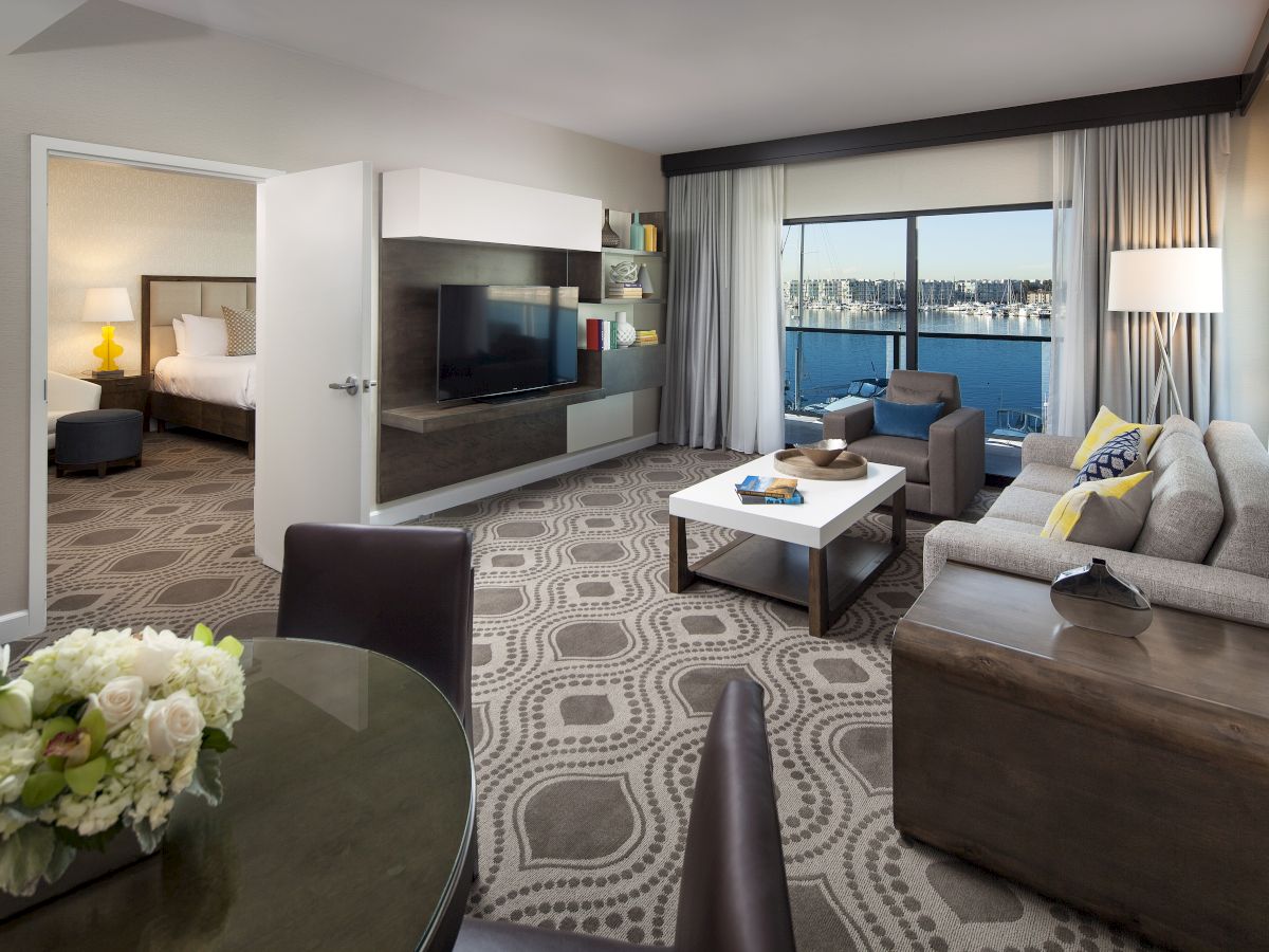 A modern hotel suite with a living area, a separate bedroom, a TV wall, a glass balcony view, and a patterned carpet.