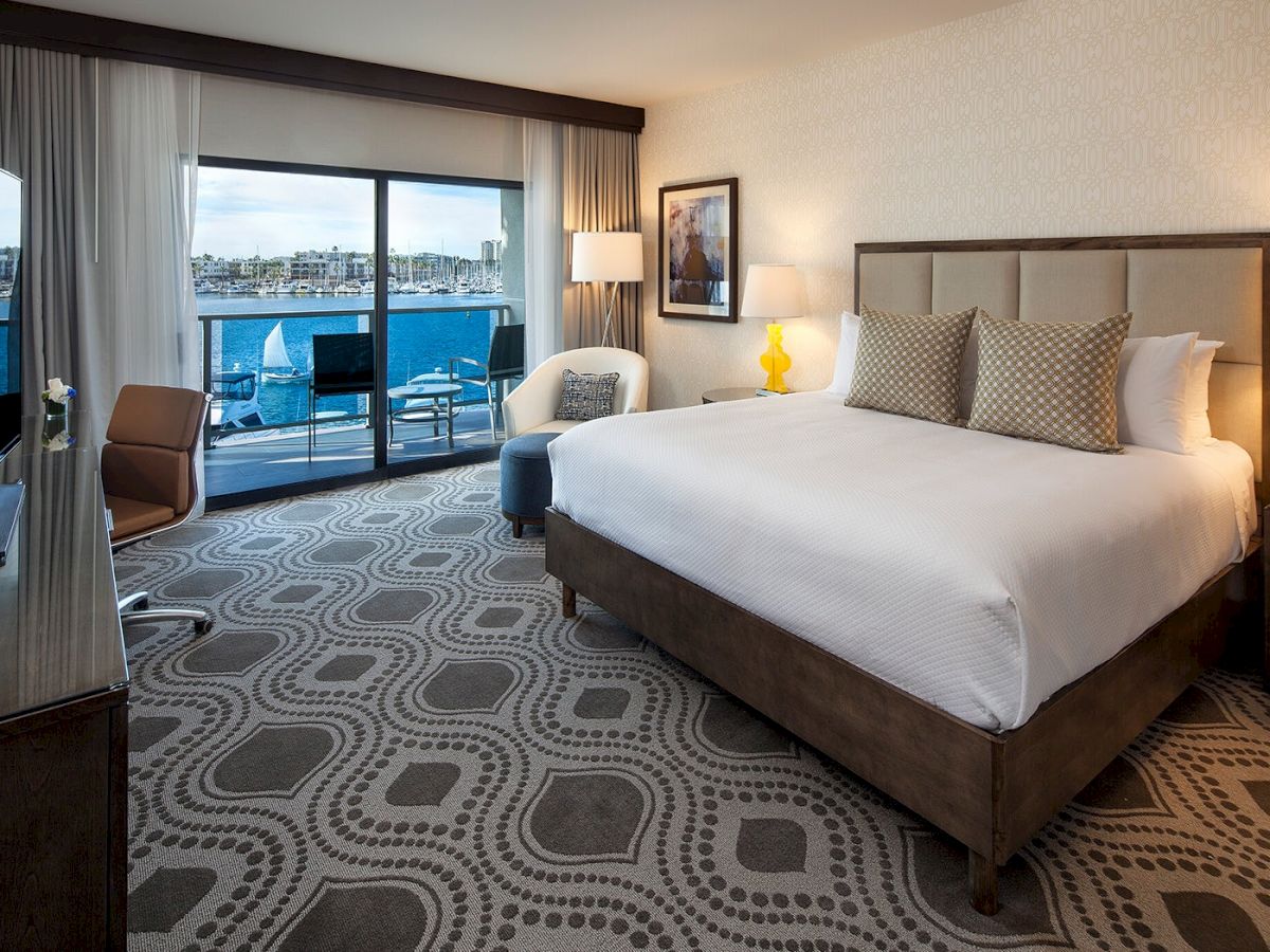 A spacious hotel room with a large bed, patterned carpet, seating area, and balcony overlooking water.