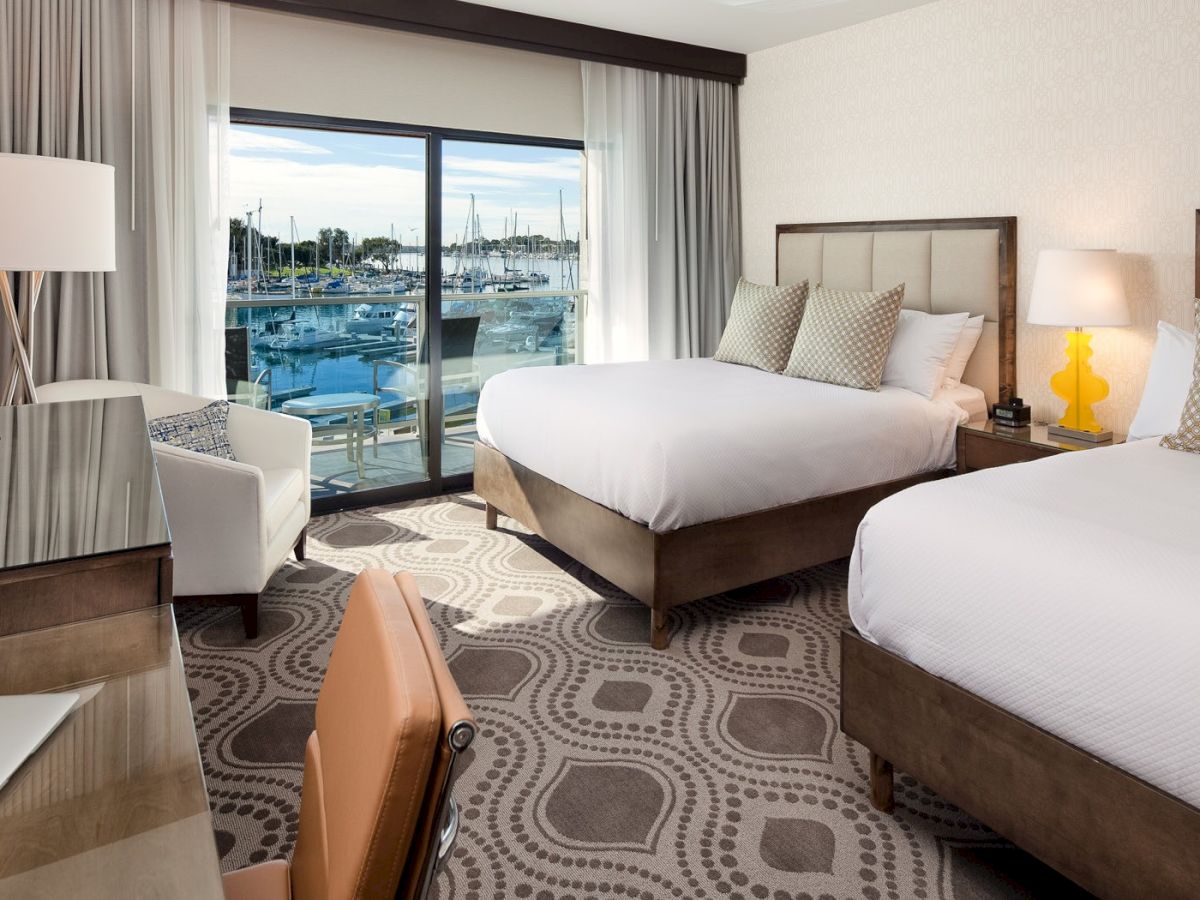 A hotel room with two beds, a desk, a chair, and a view of the water through a sliding glass door, bright and modern.