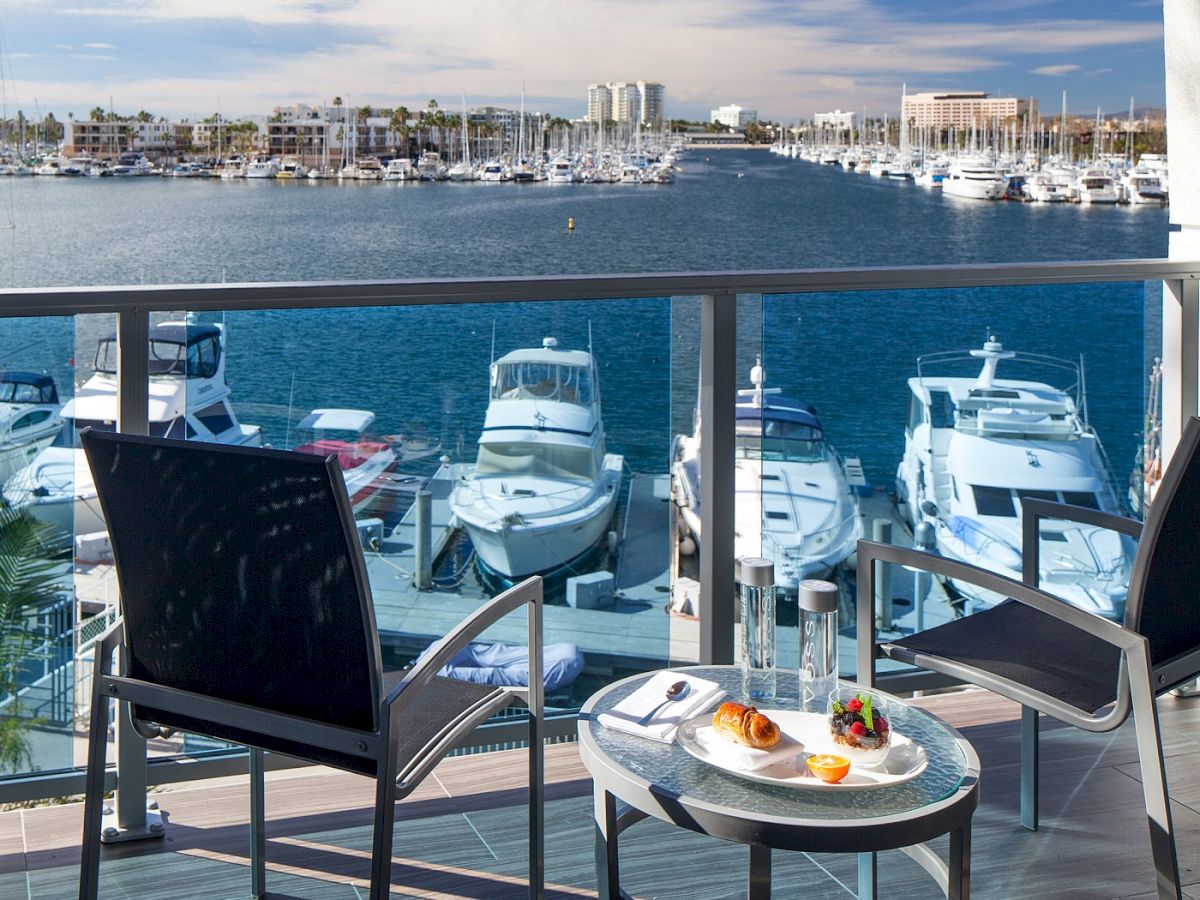 A balcony with two chairs and a small table overlooking a marina full of boats, sunny sky, and waterfront buildings in the distance.