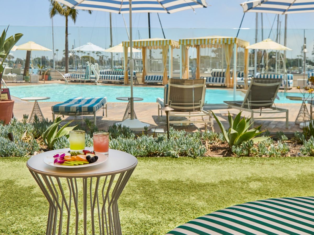 A poolside resort scene with striped chairs, umbrellas, and a white table set with drinks and fruit, lush plants, and sunny skies.