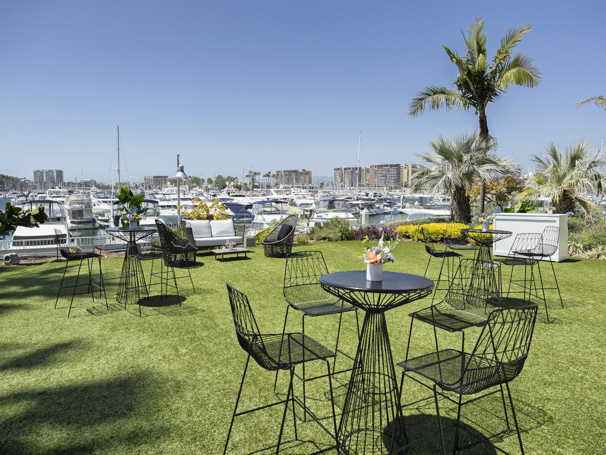 An outdoor marina cafe scene with round metal tables and chairs on a grassy lawn, palm trees, boats and a harbour in the background, bright day.