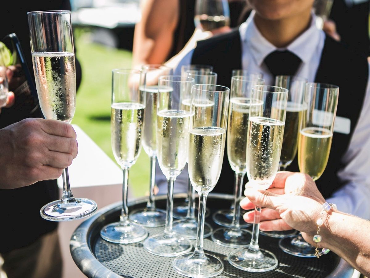 A group of people toasting with tall champagne flutes on a tray at an outdoor event, celebrating a moment together.