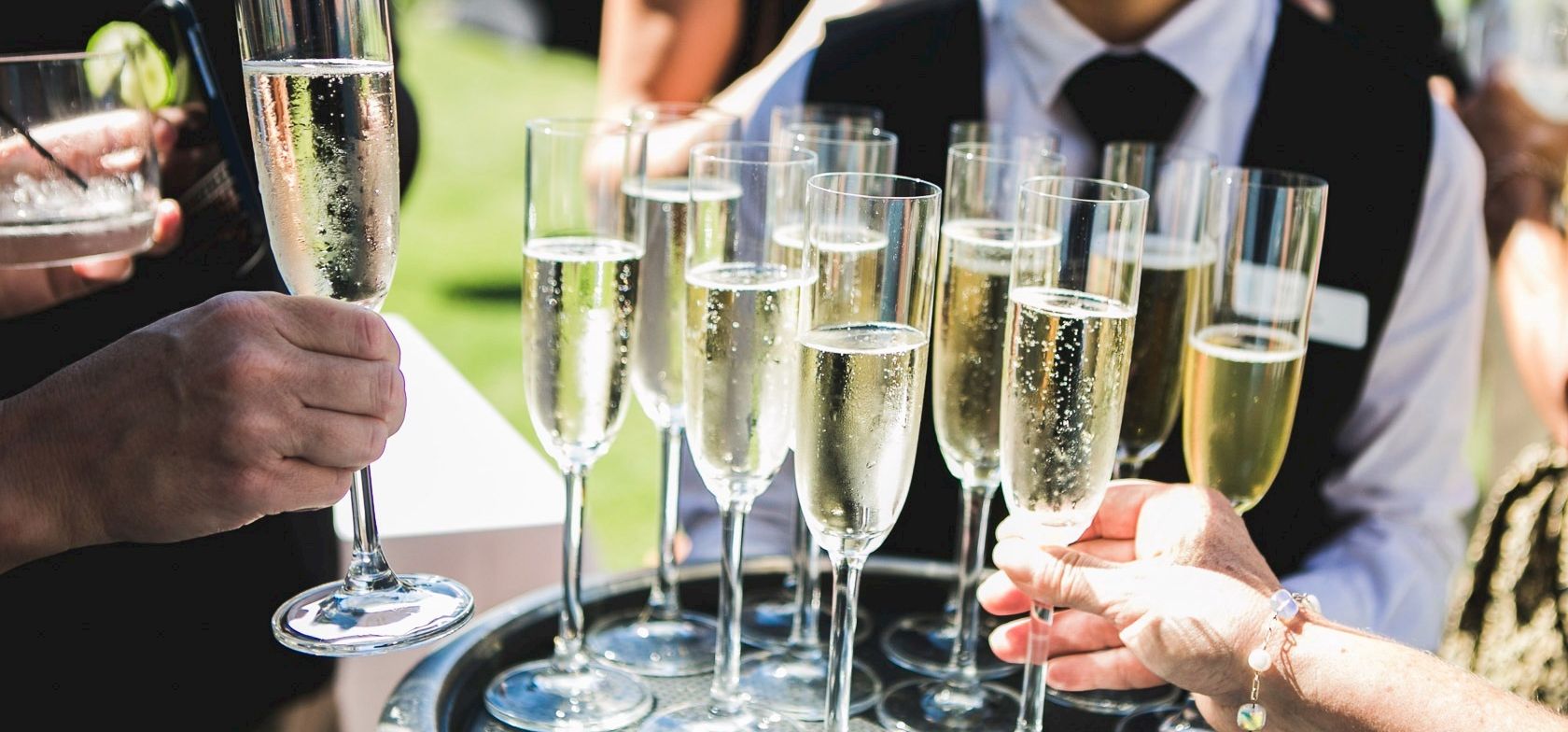 A group of people toasting with tall champagne flutes on a tray at an outdoor event, celebrating a moment together.