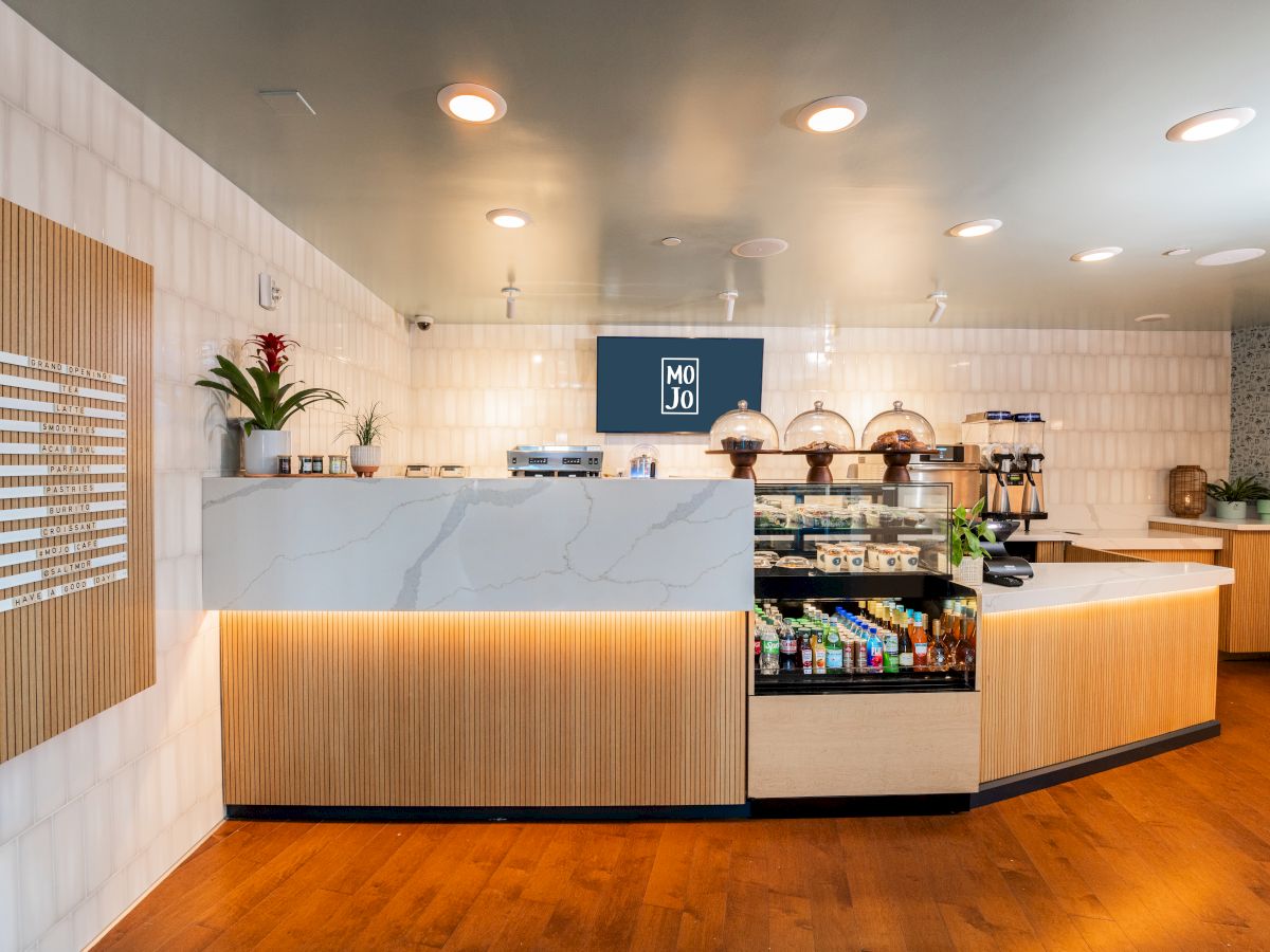 A modern cafe or bakery counter with a marble-top island, warm wood panels, display case of snacks, and a bright, welcoming interior.