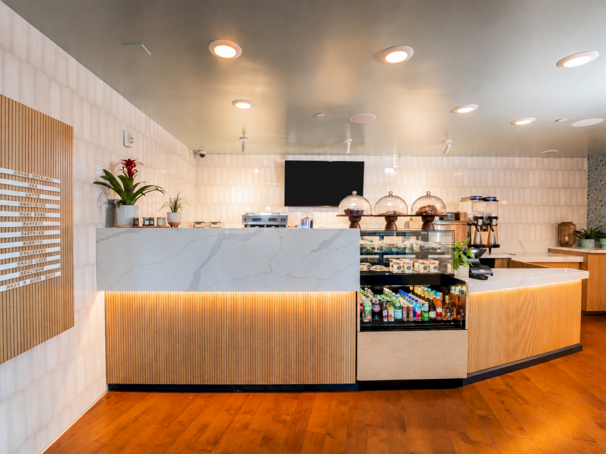 A modern caf&eacute; counter with coffee machines, pastries and drinks display, plants, and a menu sign listing items.