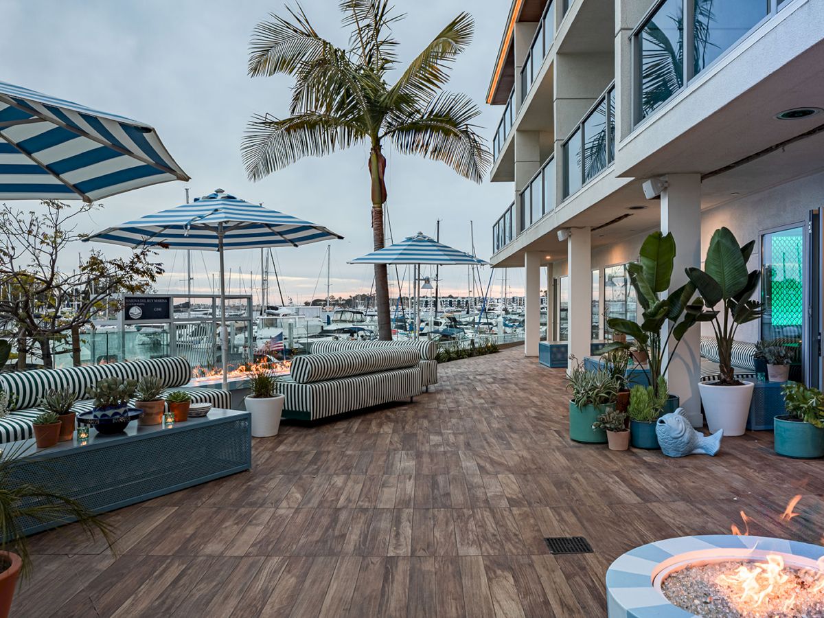 A wooden deck with potted plants, blue umbrellas, and waterfront seating along a marina, next to a modern building with balconies and a fire pit.