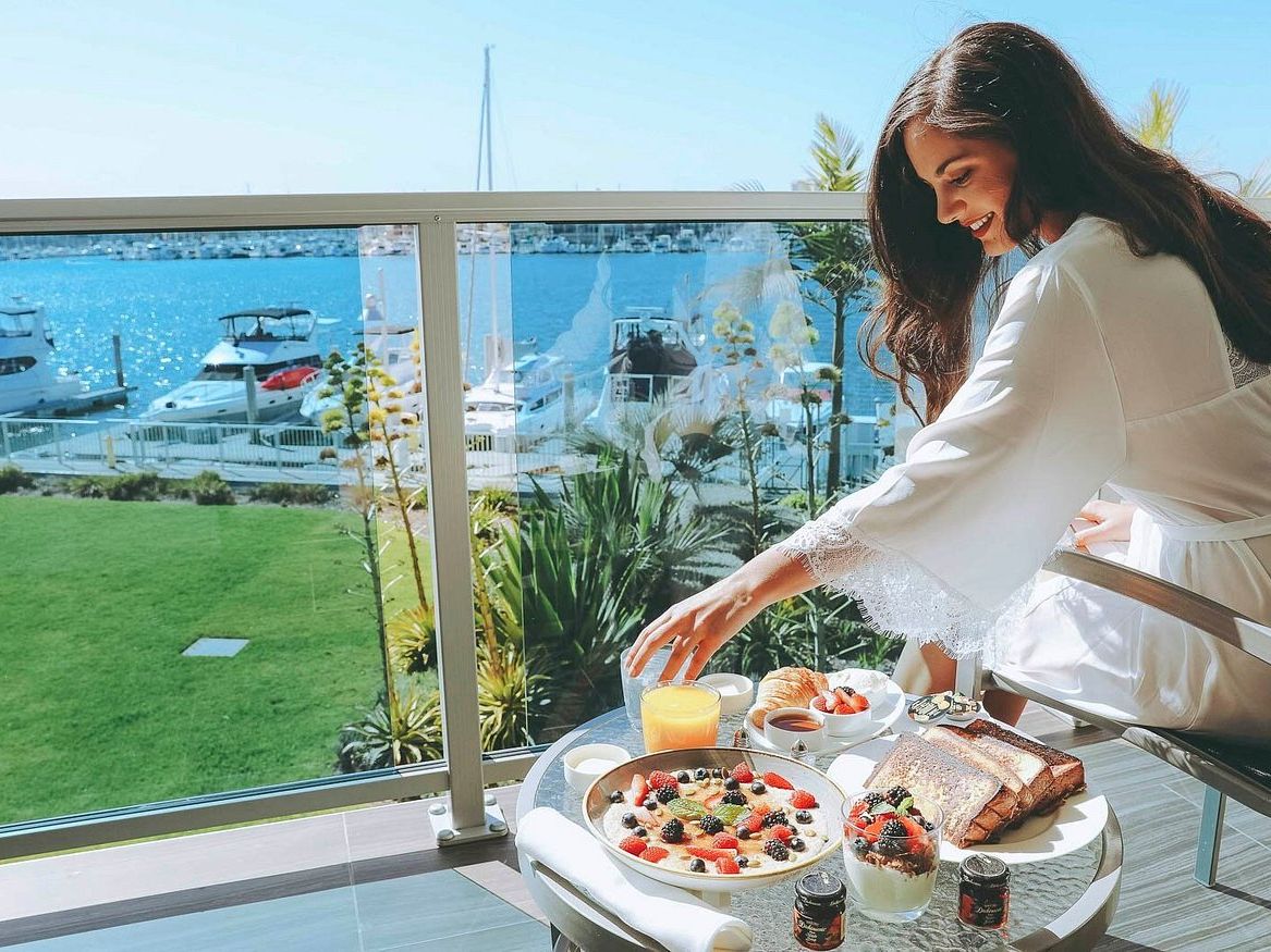 A woman enjoys a luxurious seaside breakfast on a balcony, with coffee, fruit, pastries, and a scenic marina view.