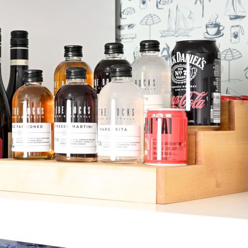 Assorted liquor bottles on a shelf with a wooden tray, including whiskey, rum, and small spirits, plus a Coca-Cola can and a white storage unit.