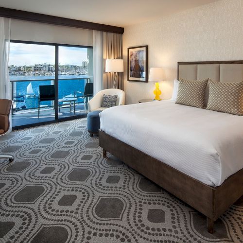 A modern hotel room with a large bed, a flat-screen TV, desk, patterned carpet, and a seating area by a window.
