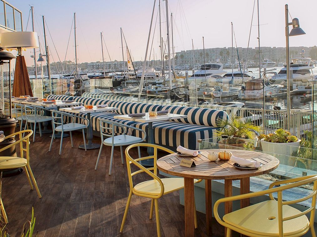 A stylish seaside restaurant deck with striped couches, round tables, yellow chairs, and boats docked in a marina in the background.