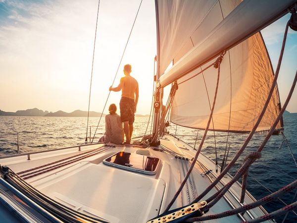 Sailing on a sailboat at sunset, two people stand near the mast on a bright deck, ropes and sails catch the wind, water glows orange.