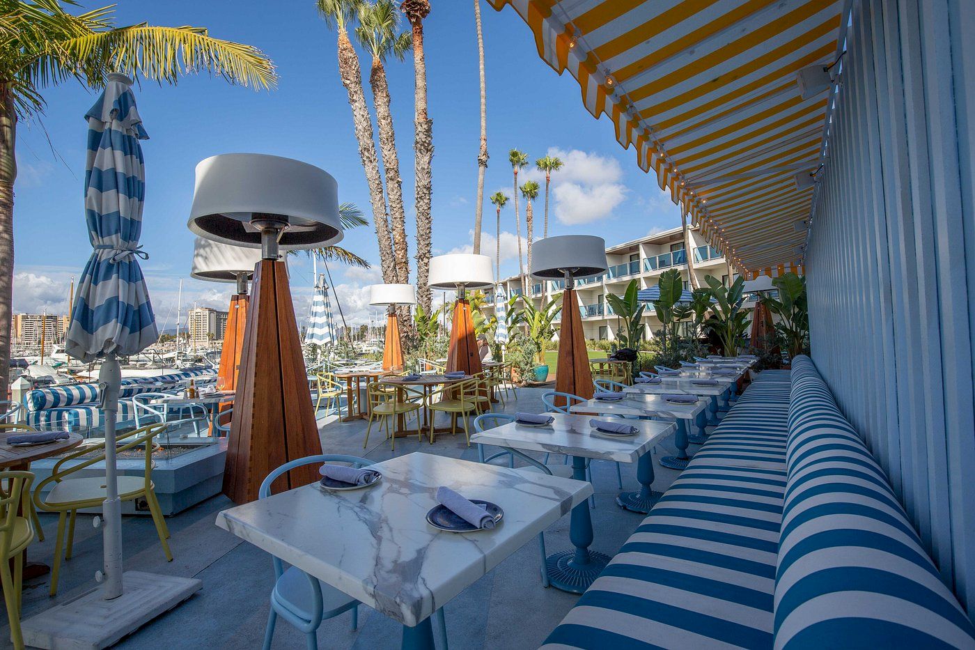 A sunny waterfront dining setup with blue-and-white striped decor, palm trees, and modern outdoor tables under shade.