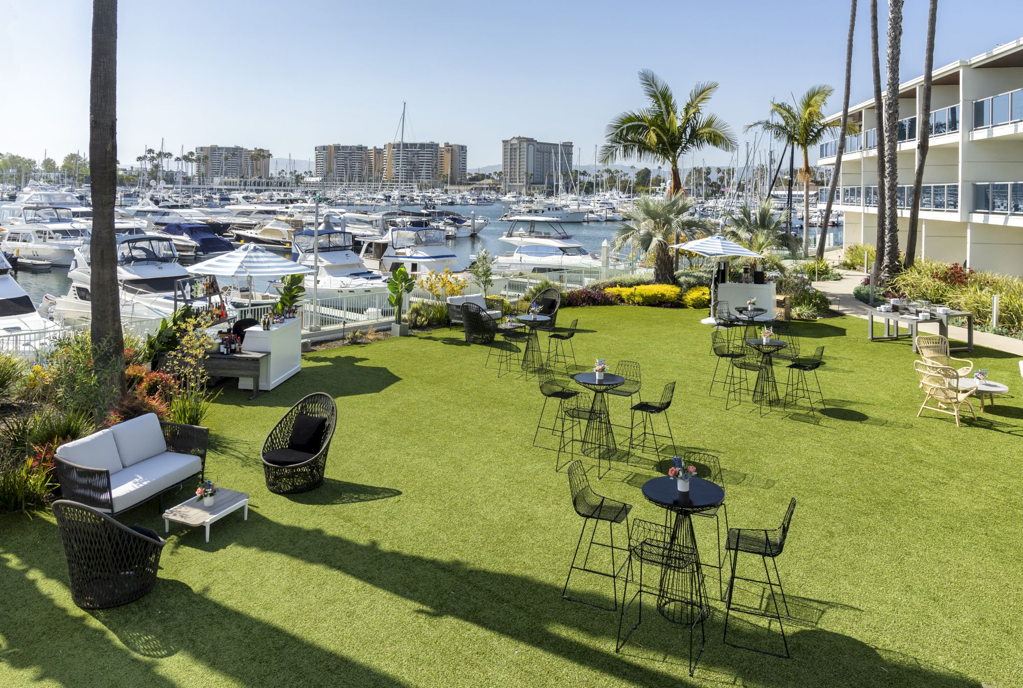 A sunny marina lawn with lounge chairs, small tables, and high-top tables, overlooking boats and palm trees by the water, perfect for relaxing.