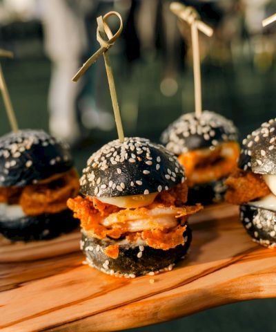 Mini sliders with black sesame buns, fried chicken, and toppings on a wooden board, each secured with a toothpick and sesame seeds.