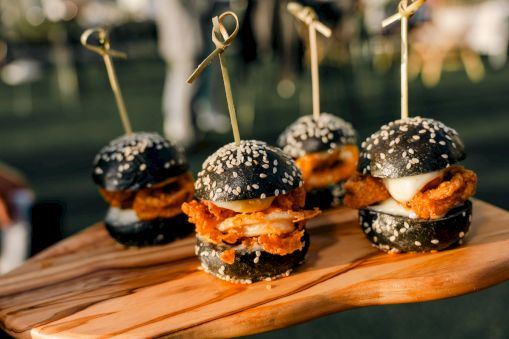 Mini sliders with black sesame buns, fried chicken, and toppings on a wooden board, each secured with a toothpick and sesame seeds.