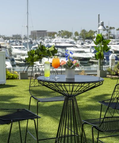 Outdoor marina cafe with black wire chairs and a round table topped by drinks and flowers, boats docked in the background by sunny water.
