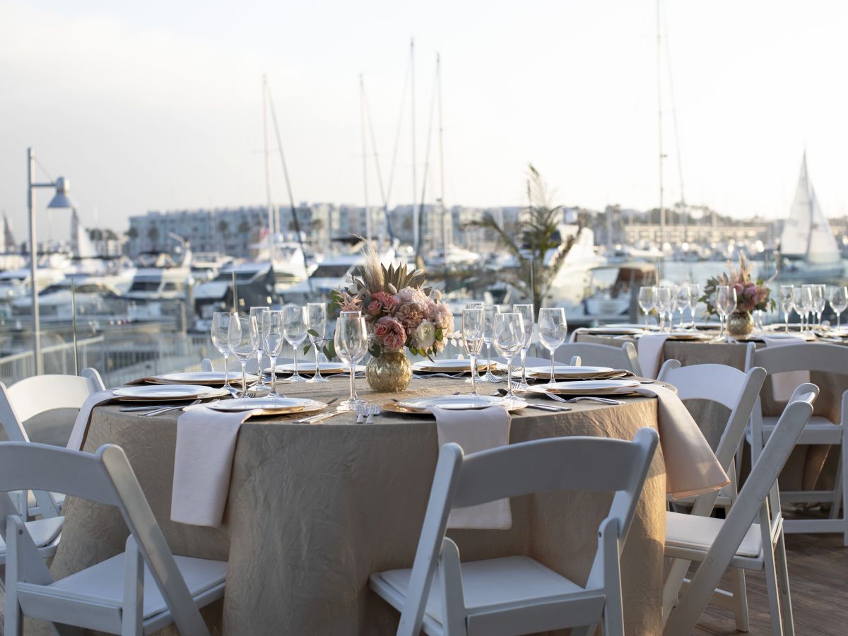 Outdoor waterfront restaurant with tables set for dining, white folding chairs, sunlit harbor, and sailboats in the background.
