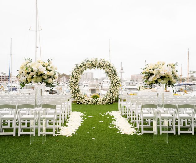 A lawn wedding setup with white chairs, floral arches and aisle petals, in front of a marina backdrop. Ends with a period.
