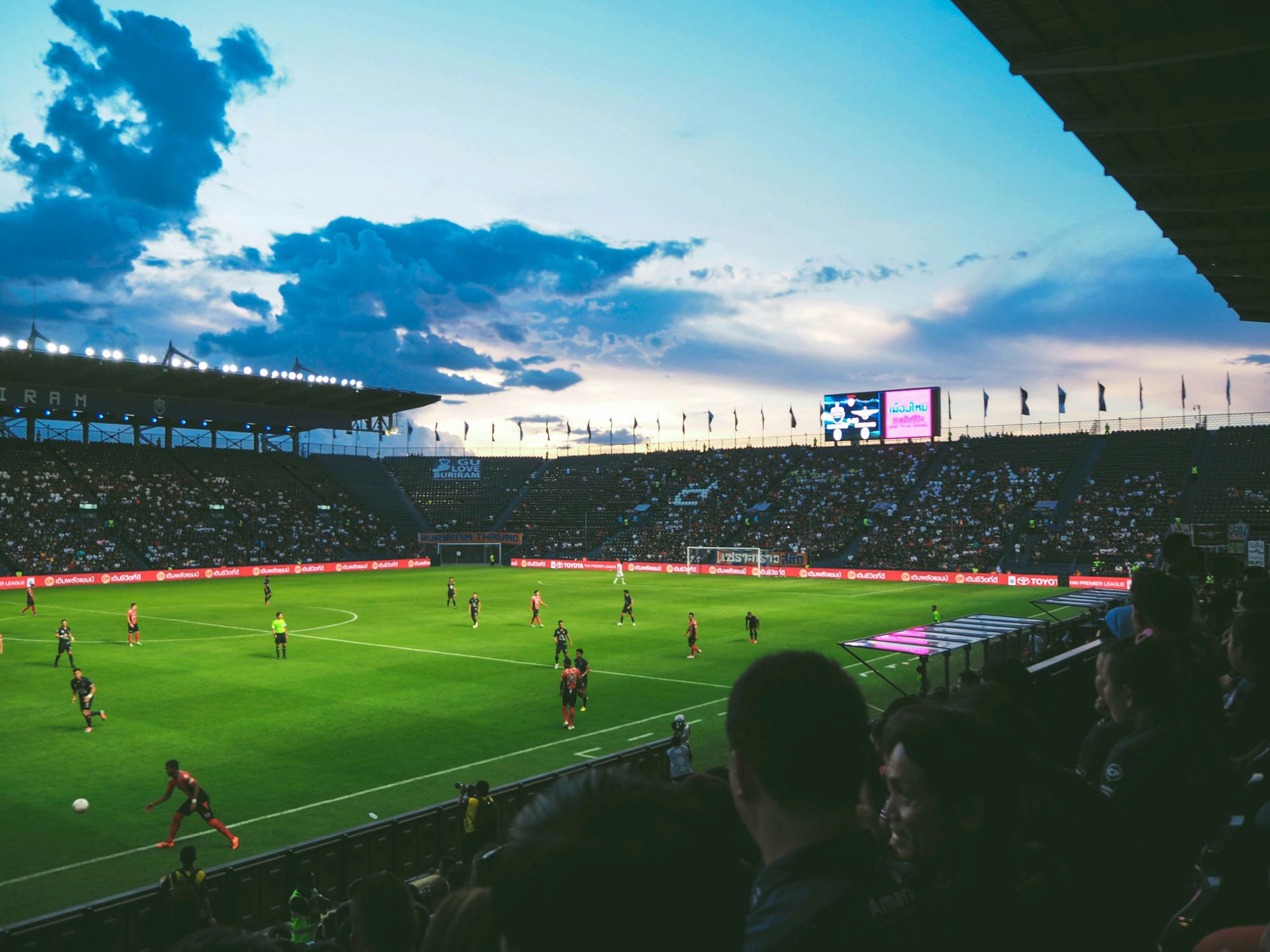 A crowded football stadium at dusk with players on the green field and spectators in the stands, under a partly cloudy sky.