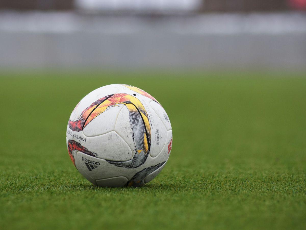 A worn soccer ball rests on a green grass field, ready for play, with a blurred stadium backdrop in the distance.
