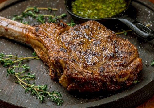 A juicy, well-seasoned bone-in rib chop on a dark plate, with herbs (thyme) and a small cup of green chimichurri or sauce in the corner.