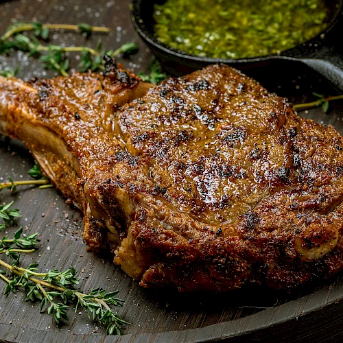 A juicy, well-seasoned bone-in rib chop on a dark plate, with herbs (thyme) and a small cup of green chimichurri or sauce in the corner.