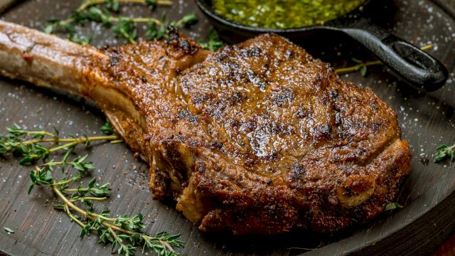 A juicy, well-seasoned bone-in rib chop on a dark plate, with herbs (thyme) and a small cup of green chimichurri or sauce in the corner.